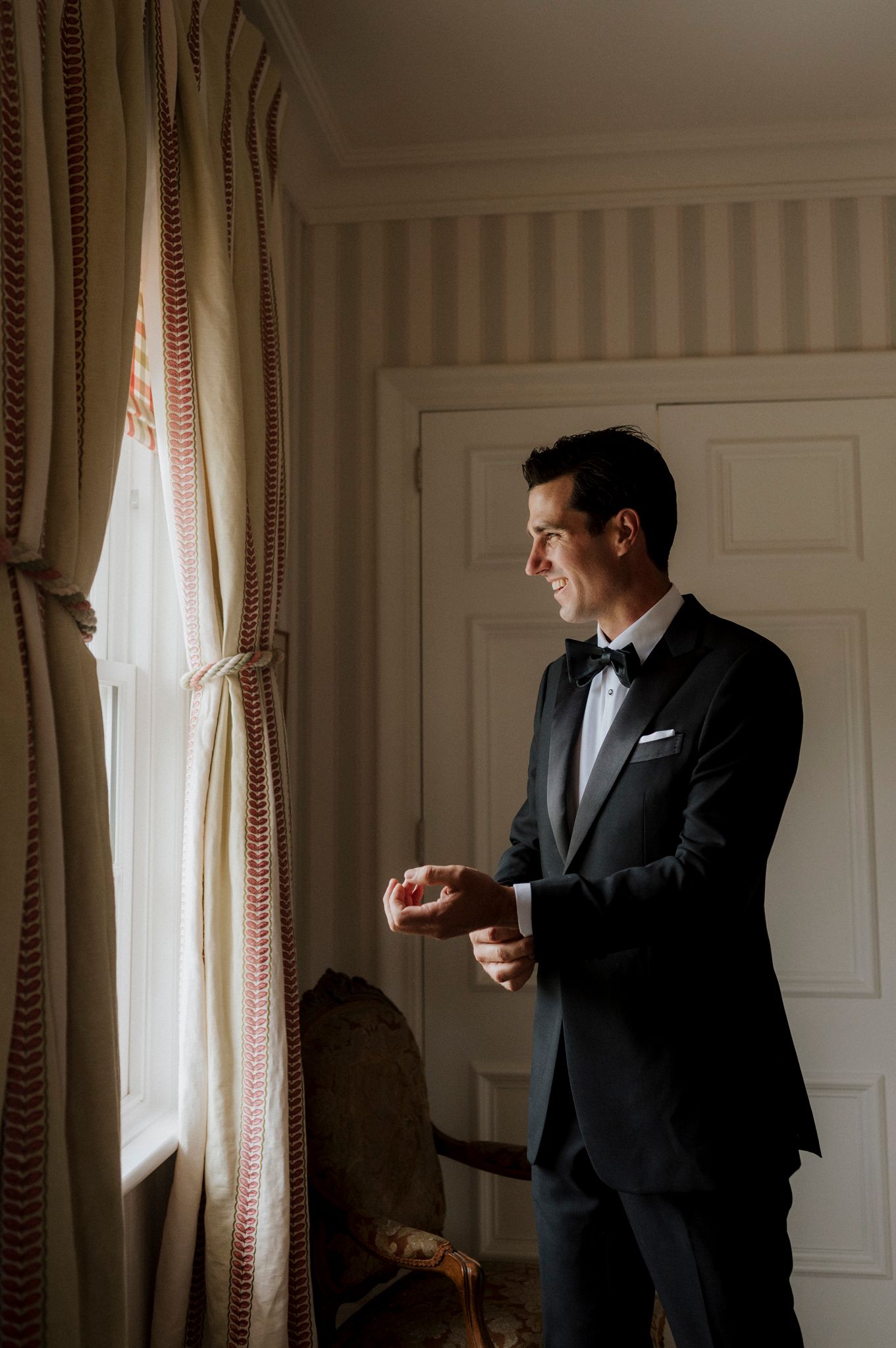 Groom in black tuxedo adjusting cufflinks by window with elegant drapes in a classic hotel room.