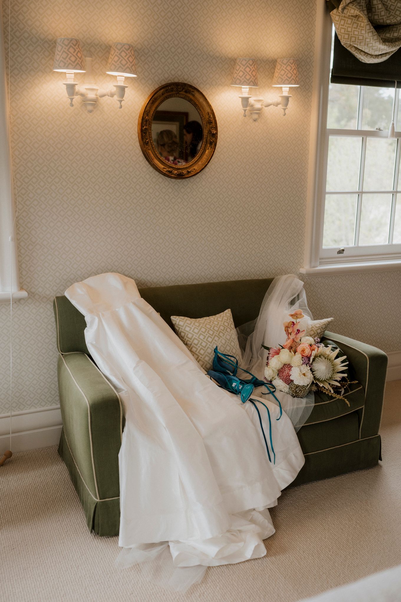 White wedding dress and bridal bouquet with pink flowers draped over a green velvet sofa in an elegant room.