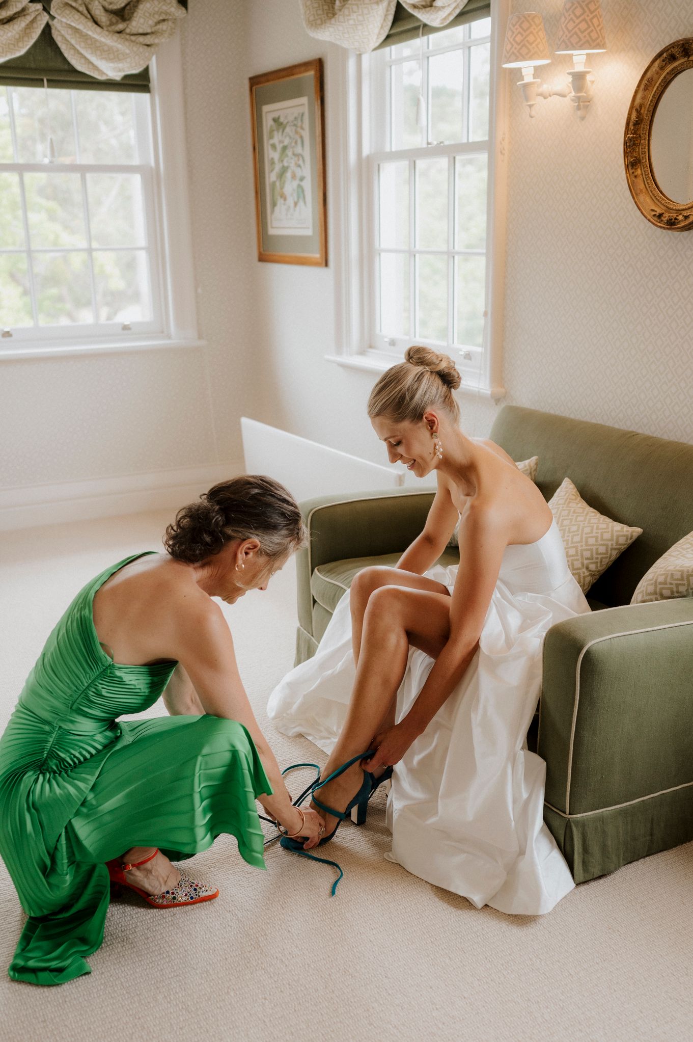 Bridesmaid in green dress helps bride put on heels before wedding ceremony in elegant room.