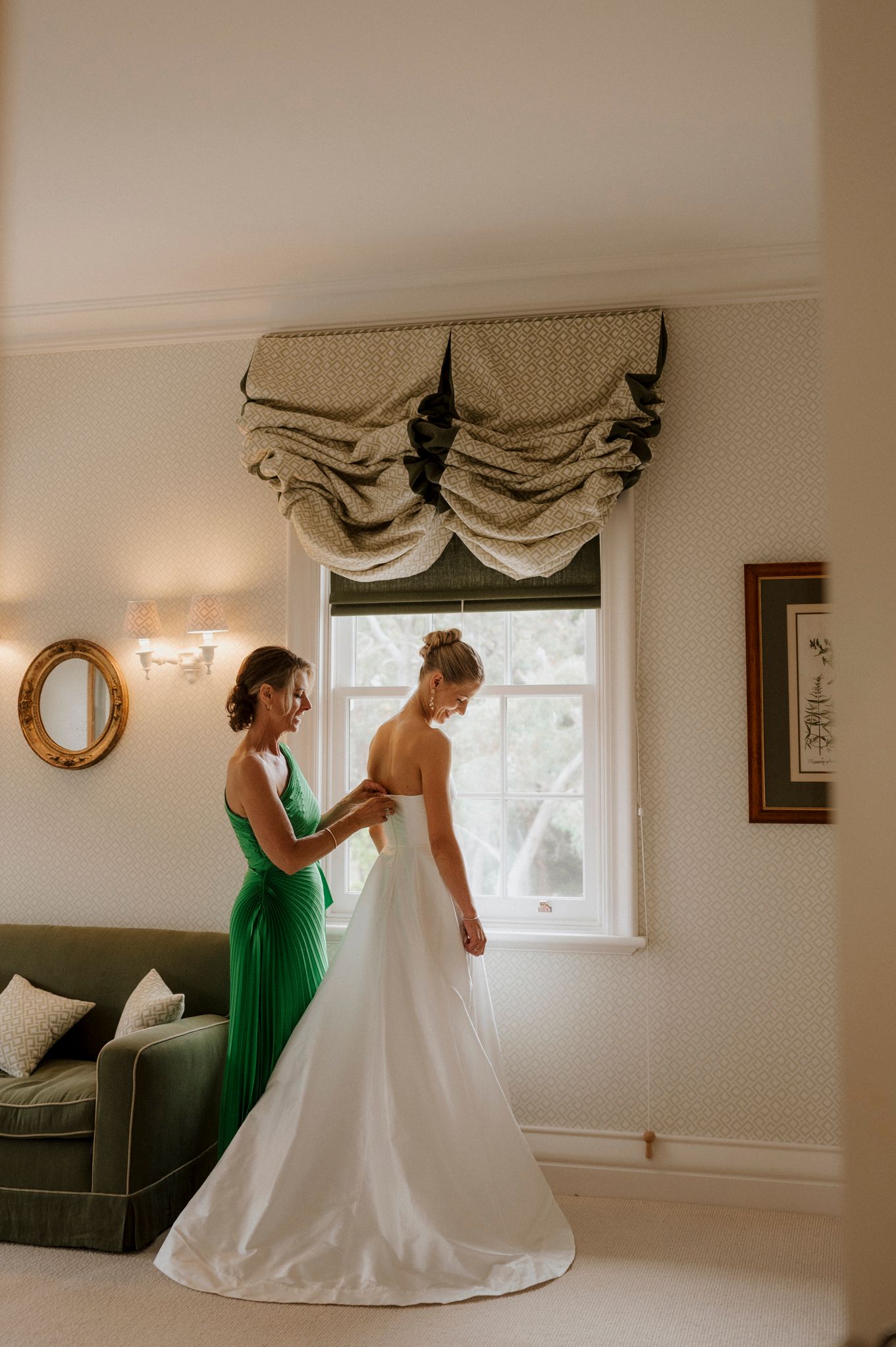 Bridesmaid in green dress helping bride with white wedding gown near window in elegant room.