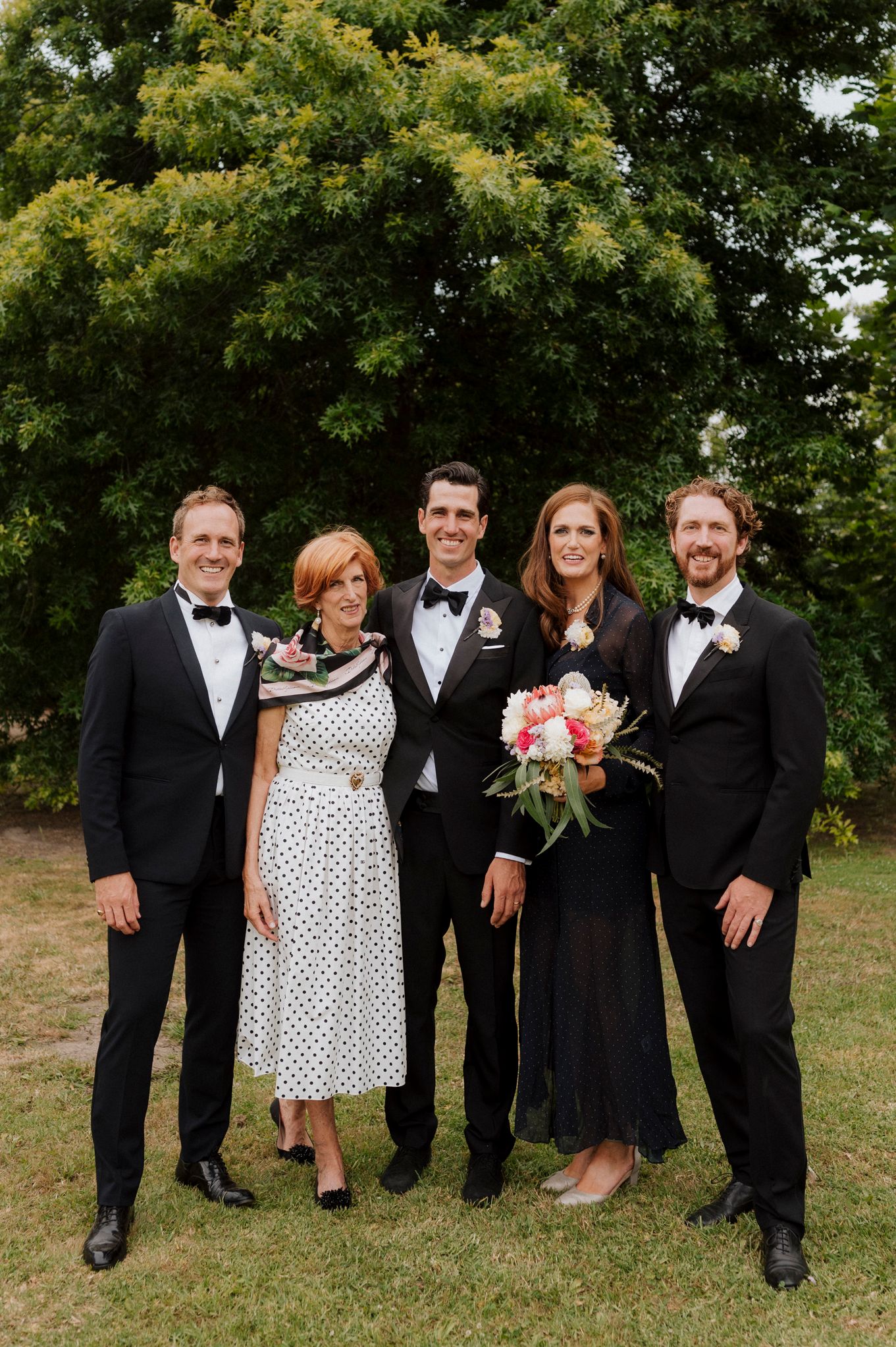 Family group photo at a wedding, with two men in tuxedos, a bride holding a bouquet, and guests posing outdoors.