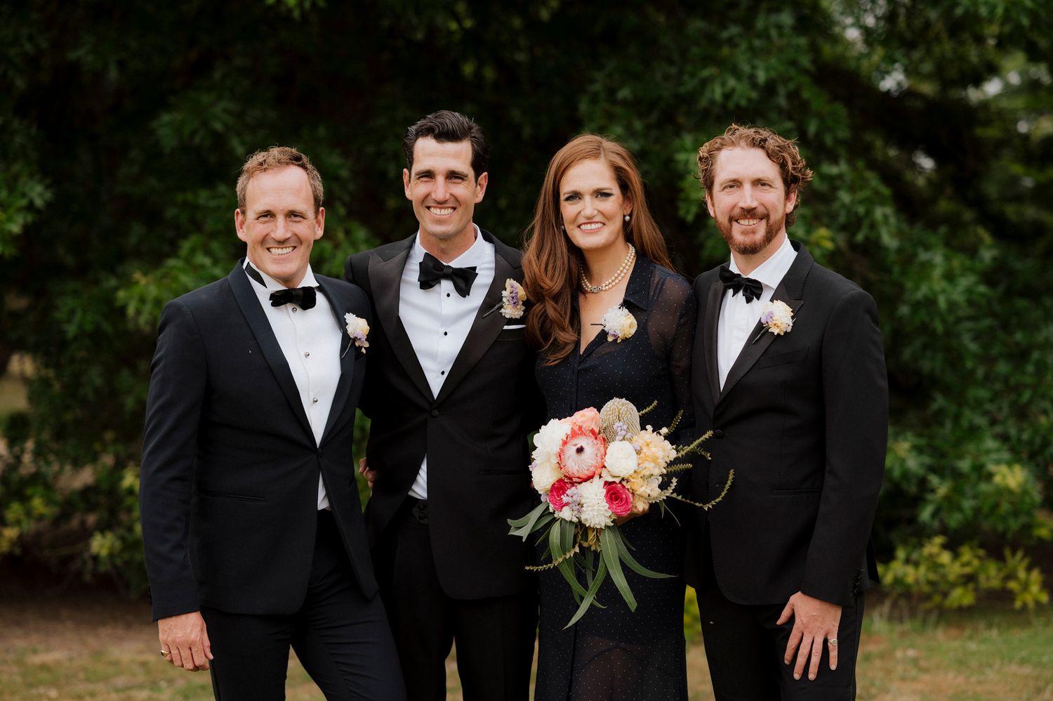 Wedding party in black tuxedos and gown posing outdoors, bride holding colorful floral bouquet.