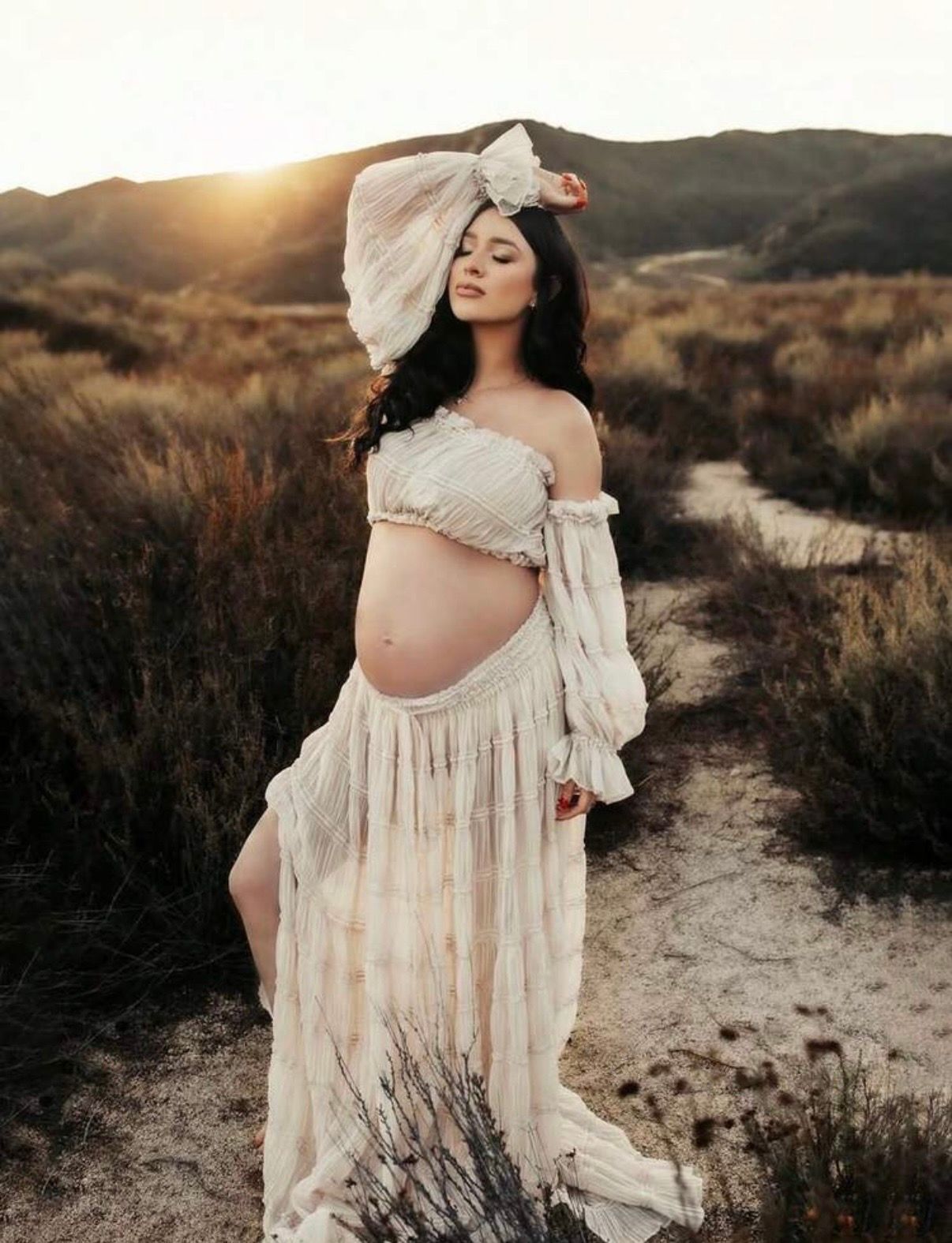 Pregnant woman in boho white dress posing outdoors at golden hour in a scenic desert landscape.