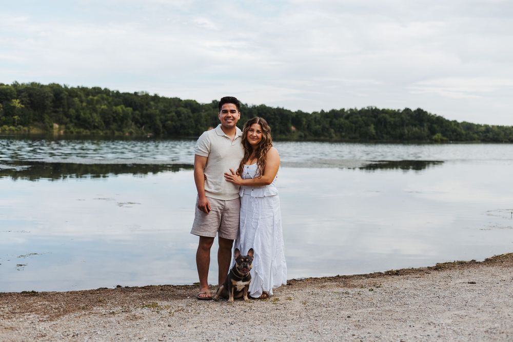 Couple and French Bulldog posing on a lakeside beach with lush green trees in the background.