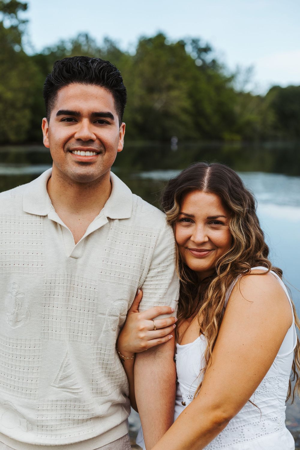 A smiling couple poses together near a lake with trees in the background on a warm evening.