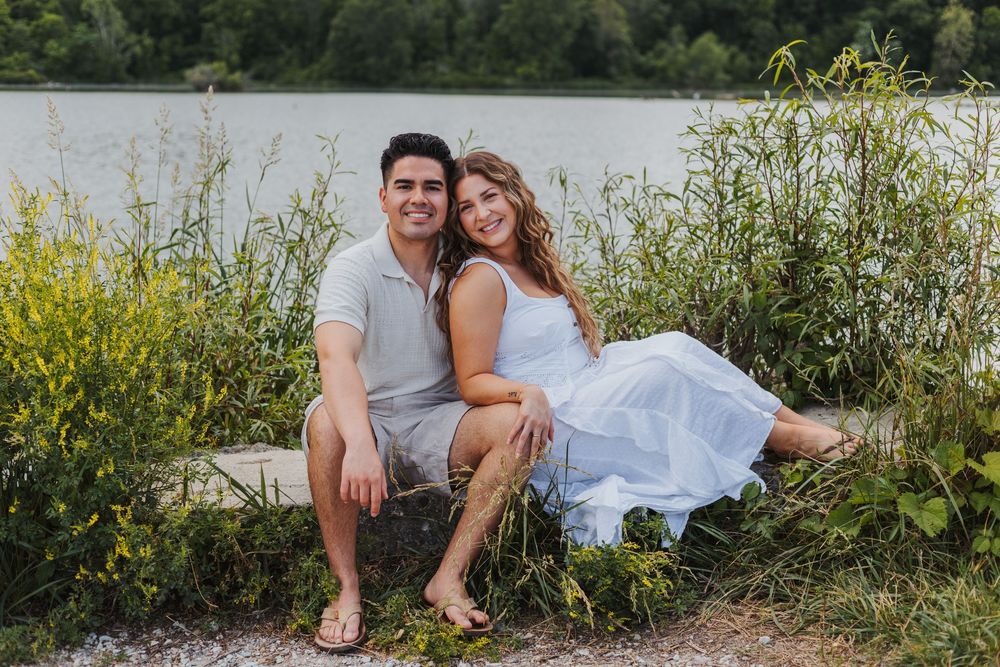 A smiling couple sits together on a rock near a lake surrounded by lush green plants on a sunny day.