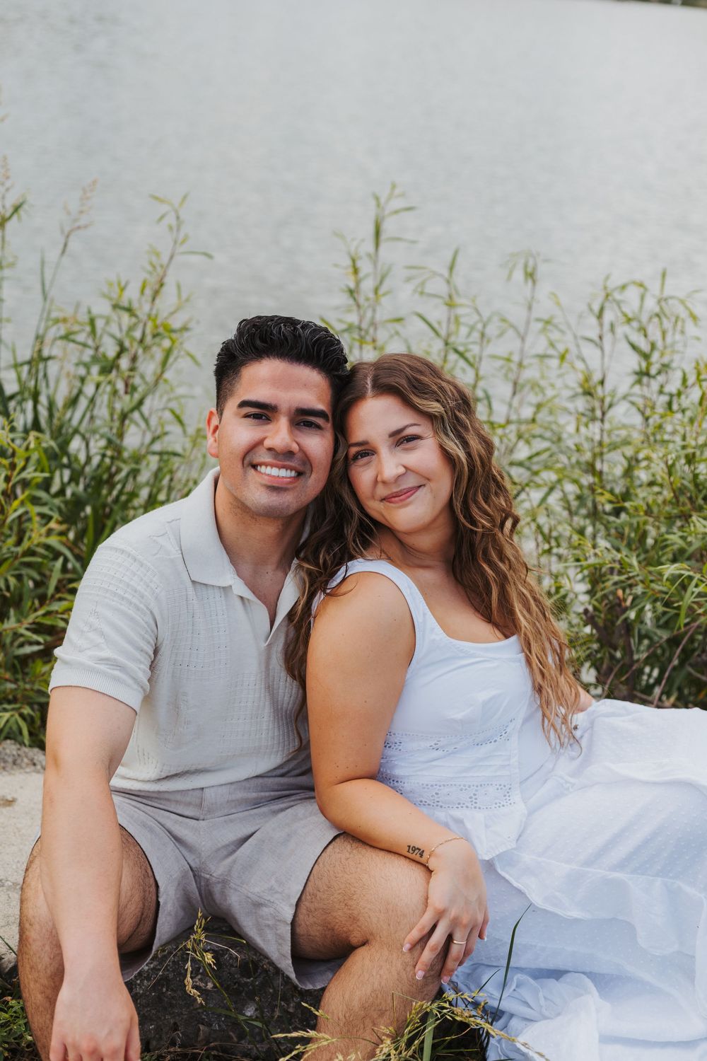 A smiling couple sits together by a serene lakeside with lush green reeds, wearing coordinated light blue outfits.