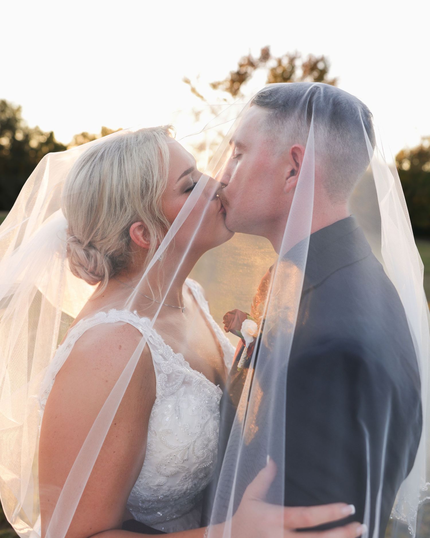 Bride and groom sharing a romantic kiss under a flowing white veil at golden hour outdoors.
