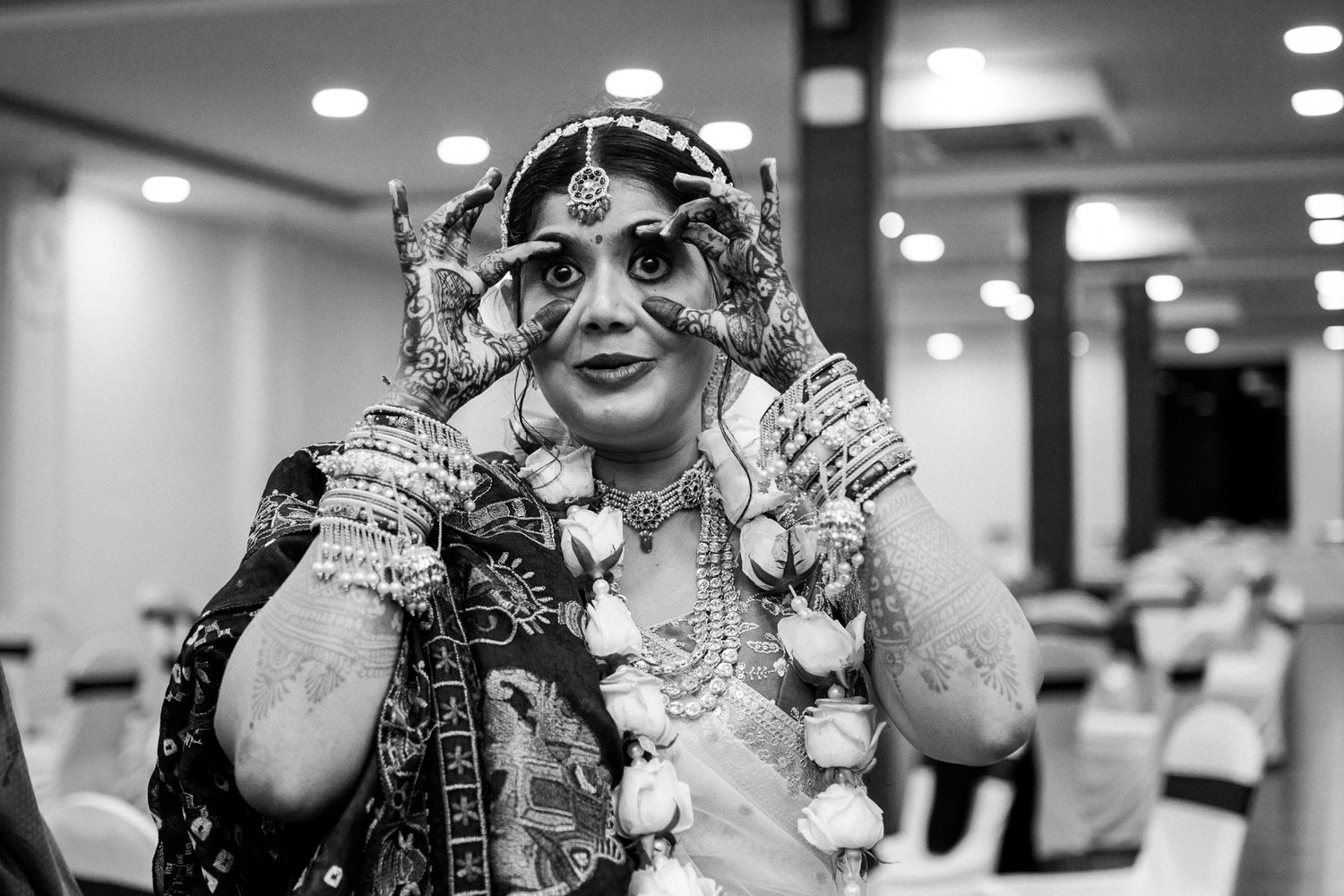 Black and white photo of an Indian bride in traditional jewelry and bridal attire adjusting her accessories indoors.