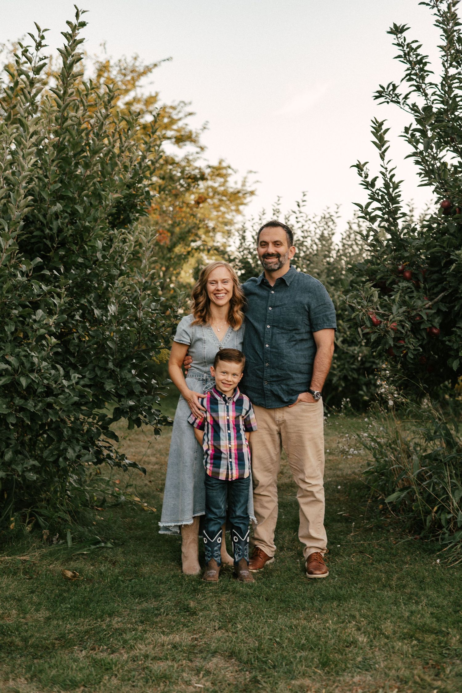 Family of three smiling in an apple orchard, parents standing with young boy in plaid shirt and cowboy boots.