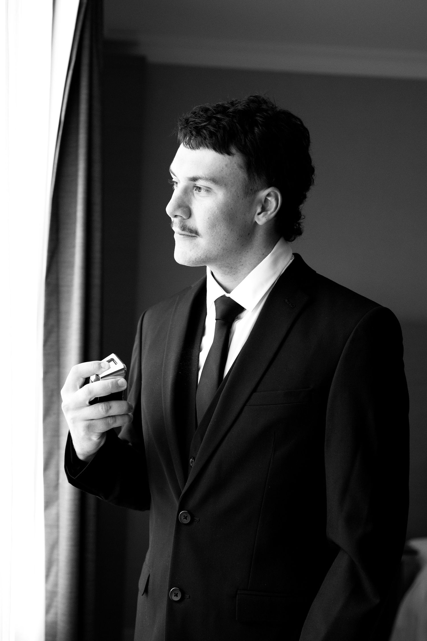 Black and white portrait of a young man in a suit holding cufflinks, gazing thoughtfully out a window.