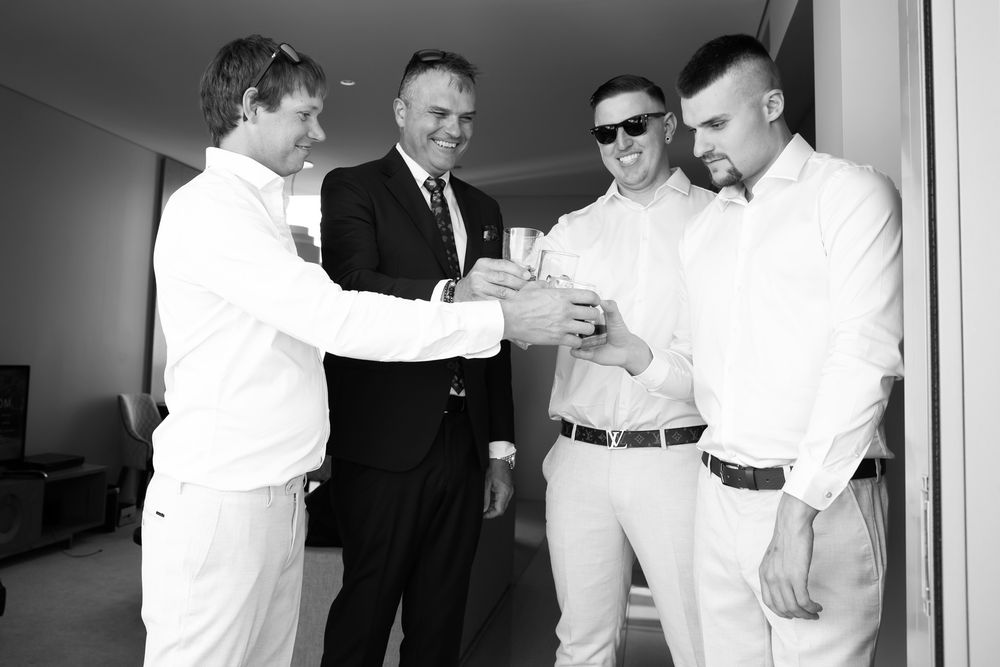 Four men in white shirts and one in a suit toasting drinks together in a bright room.