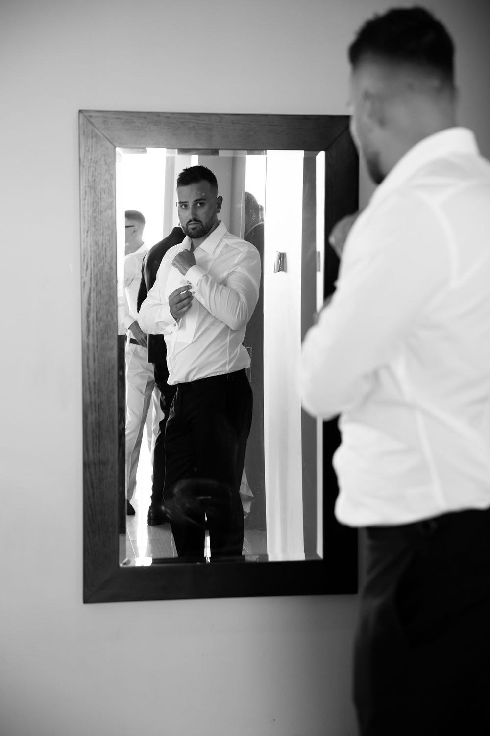 Black and white photo of a well-dressed groom adjusting his tie while looking at his reflection in a wooden framed mirror.