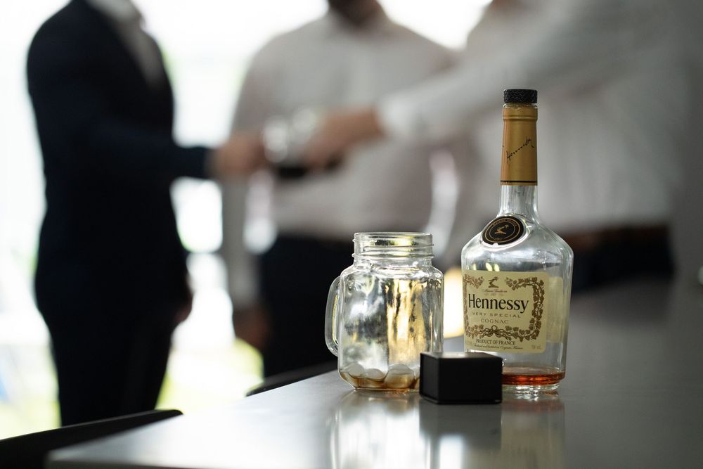 Hennessy cognac bottle on a table with a glass jar and blurred men in suits in the background.