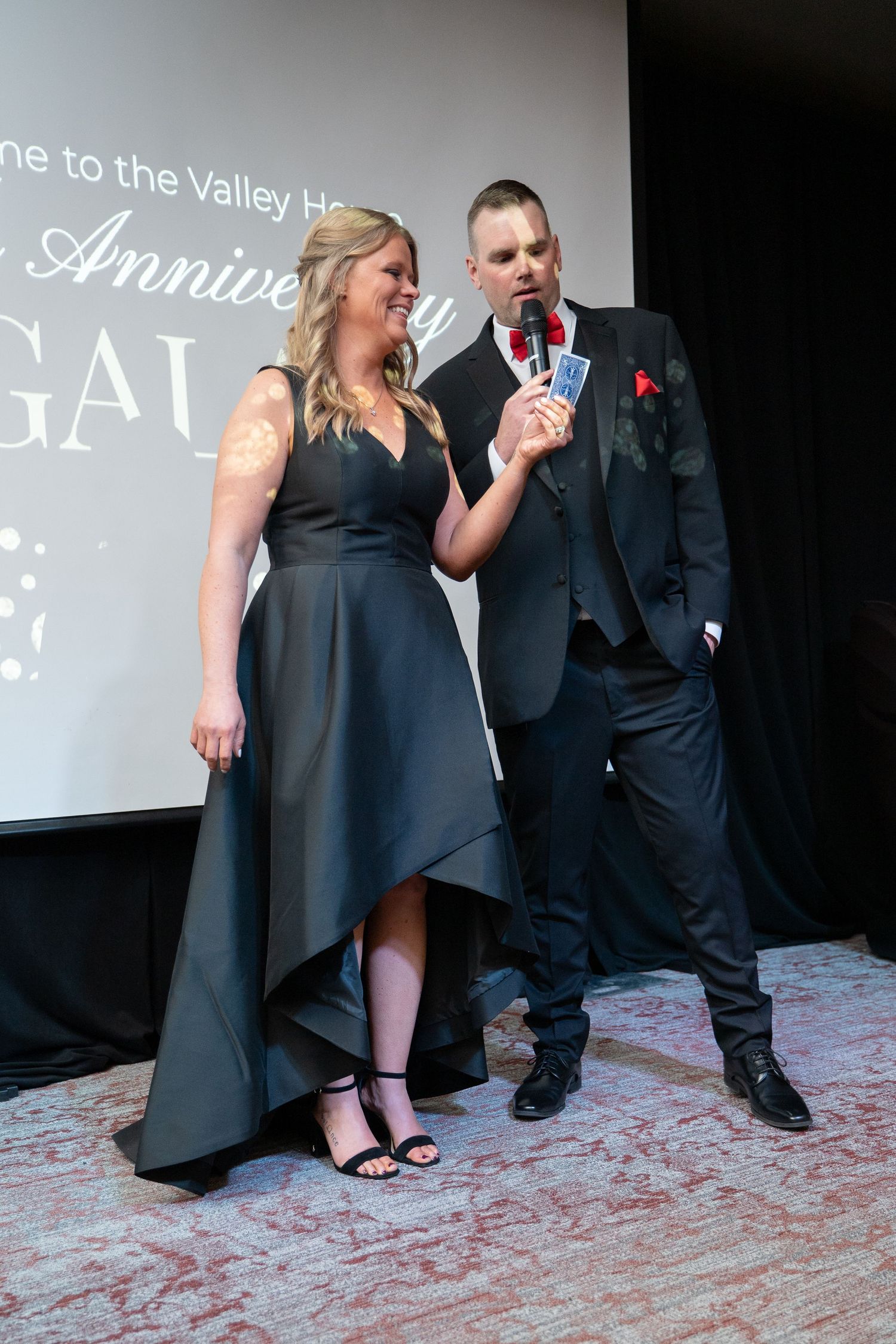 Woman in elegant black high-low dress and heels walking at a formal gala event with decorated tables.