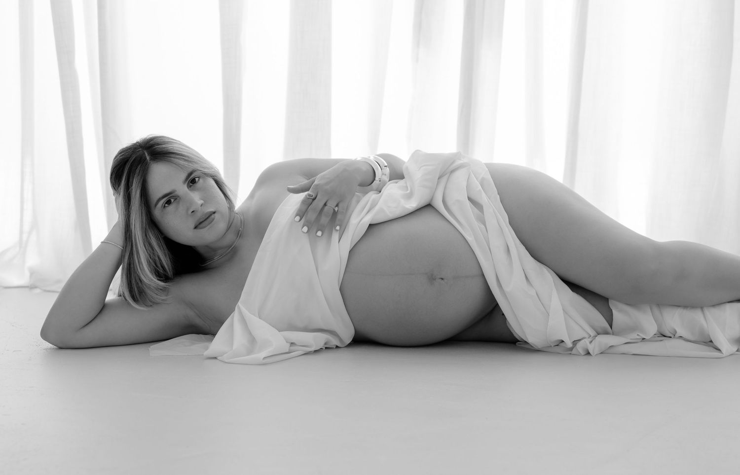 Black and white maternity photo of a pregnant woman lying down, draped in white fabric, showcasing her baby bump.