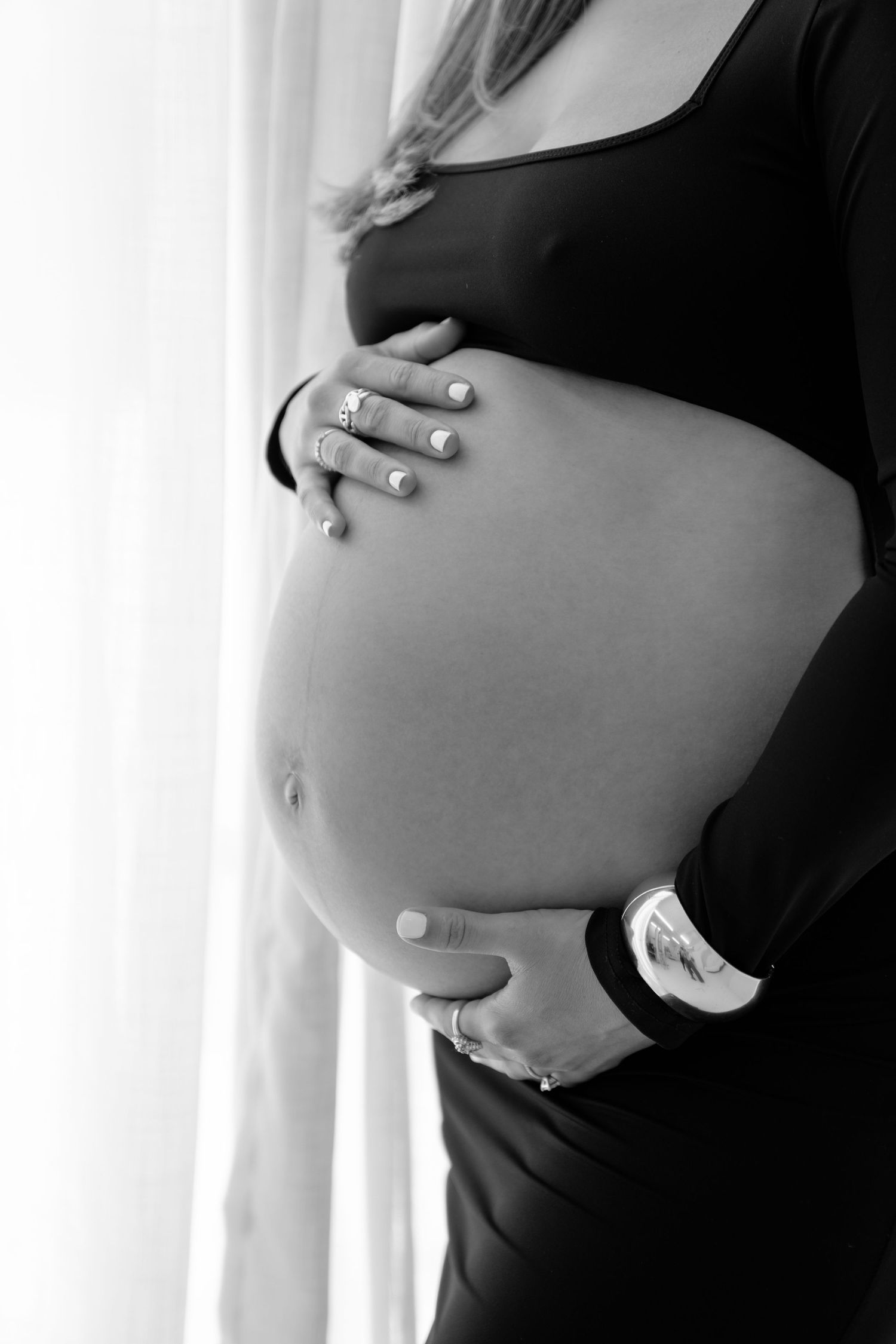 Black and white photo of a pregnant woman in a black dress cradling her baby bump near a bright window.