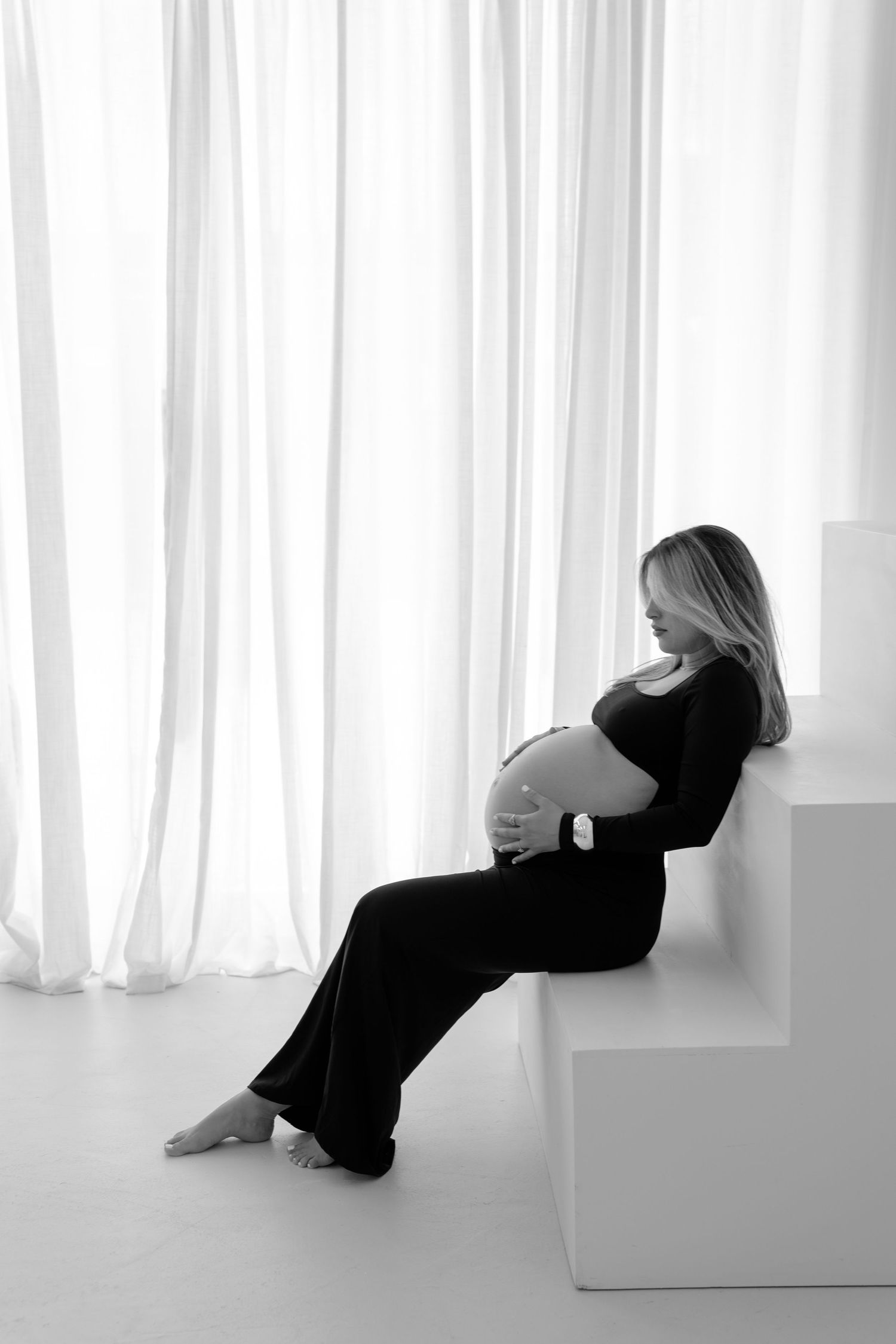 Black and white photo of a pregnant woman in a black dress sitting and cradling her baby bump near sheer white curtains.