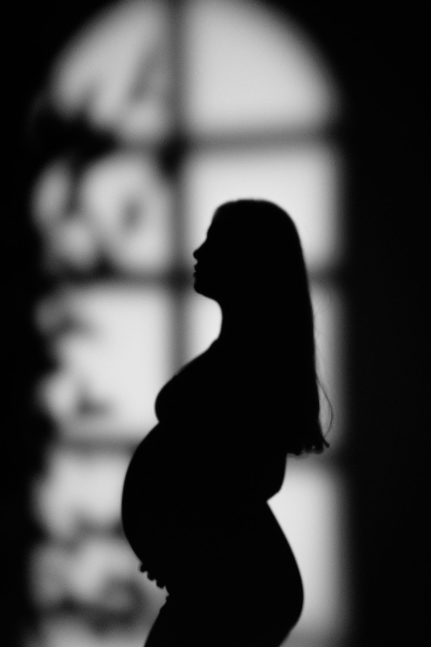 Black and white silhouette of a pregnant woman standing in front of an arched window, showcasing her baby bump.