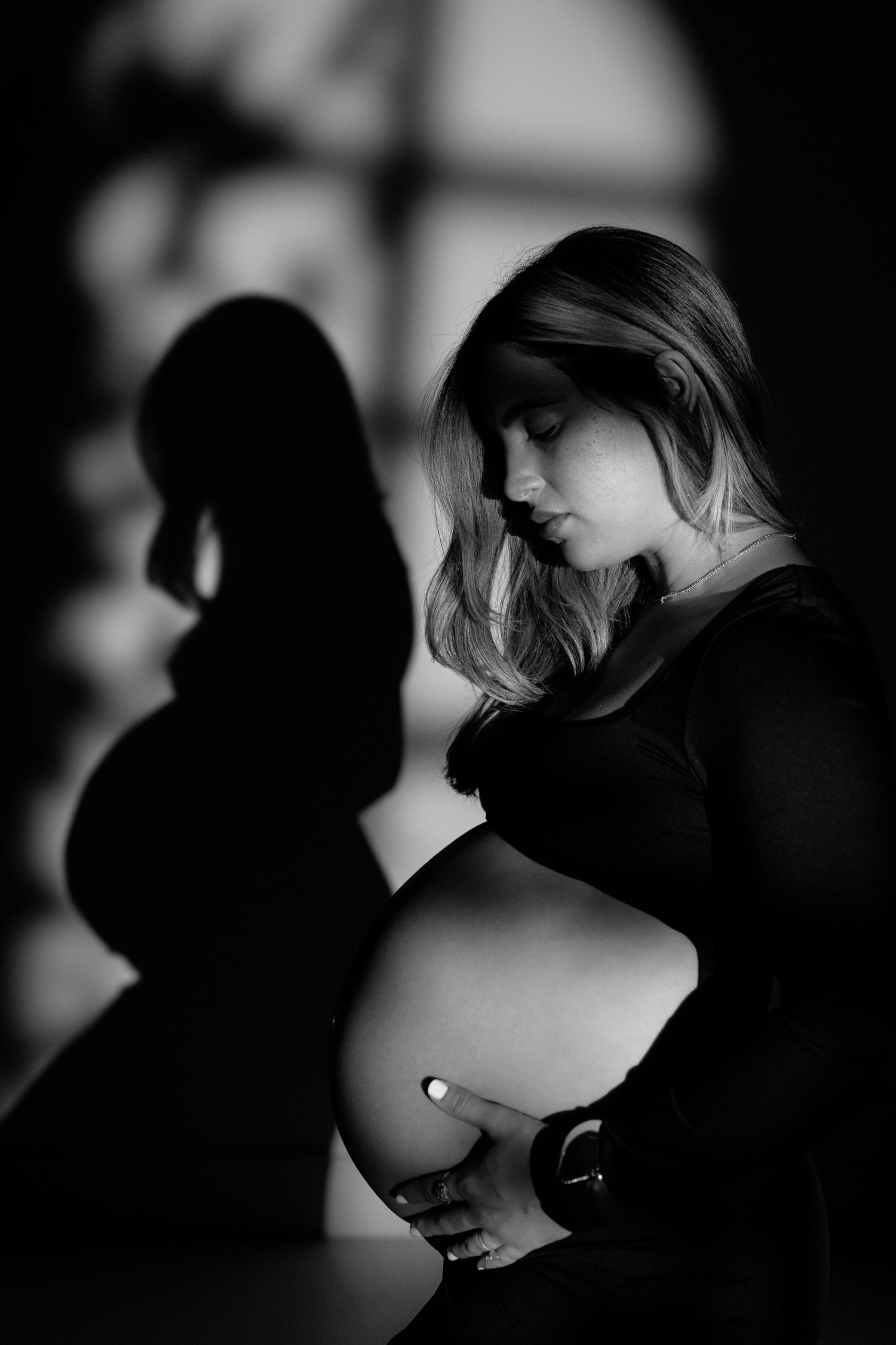 Black and white portrait of a pregnant woman cradling her baby bump with her shadow cast on the wall behind her.