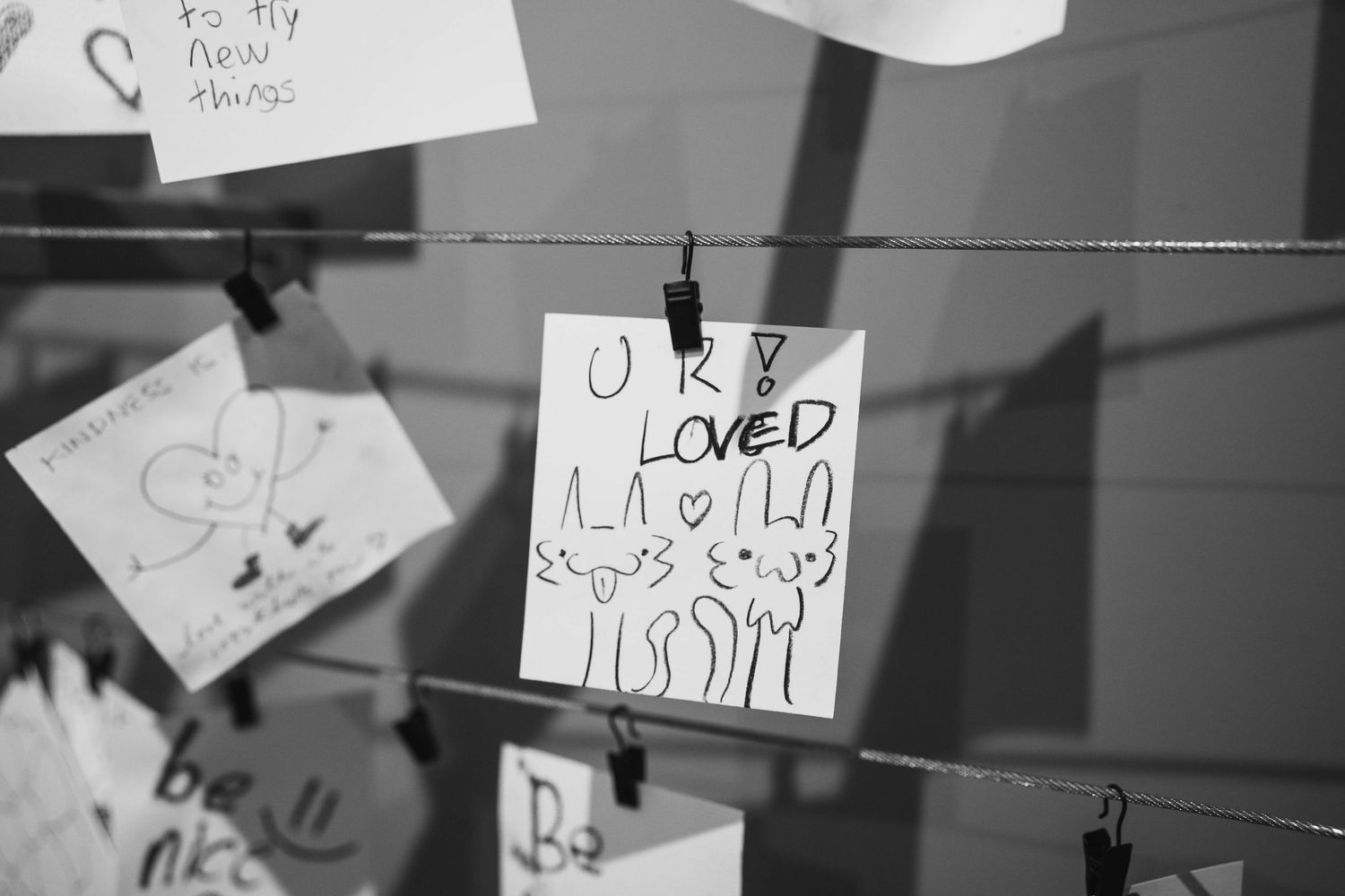 Black and white photo of handwritten notes on clips, with one card reading U R LOVED hanging on a wire display.