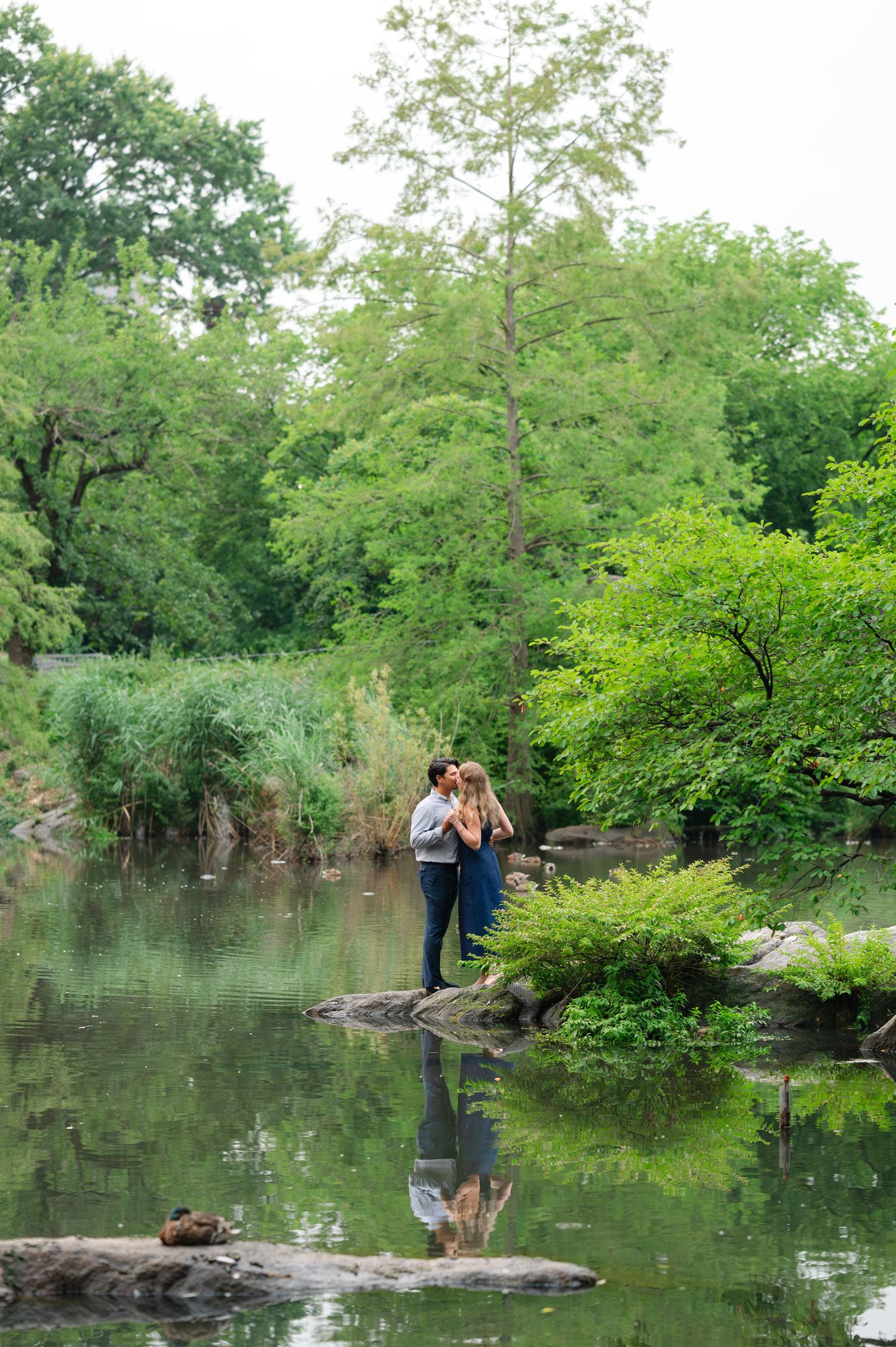 Couple sharing a romantic kiss on rocks by a serene pond surrounded by lush green trees in Central Park near the Gapstow Bridge.