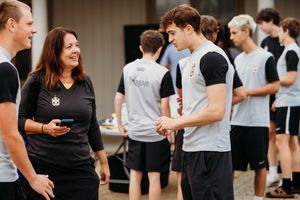 Woman in black polo shirt conversing with young male athletes outdoors at a sports training event.