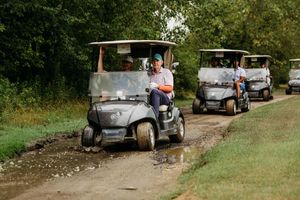 A convoy of golf carts drives along a muddy dirt path surrounded by lush green trees on a golf course.