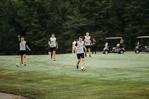 Young men playing soccer on a lush green golf course with trees and a golf cart in the background.