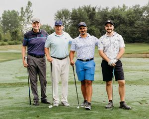 Four men holding golf clubs pose together on a golf course surrounded by lush greenery.