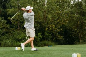 Golfer in white cap and khaki shorts swinging a golf club on a lush green golf course.