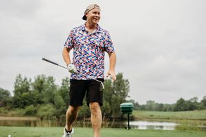 Man smiling on golf course wearing colorful patterned shirt and black shorts, holding a golf club near a pond.