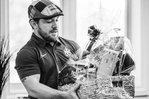 Man in cap and Nike shirt examines a large gift basket filled with various items near a bright window.