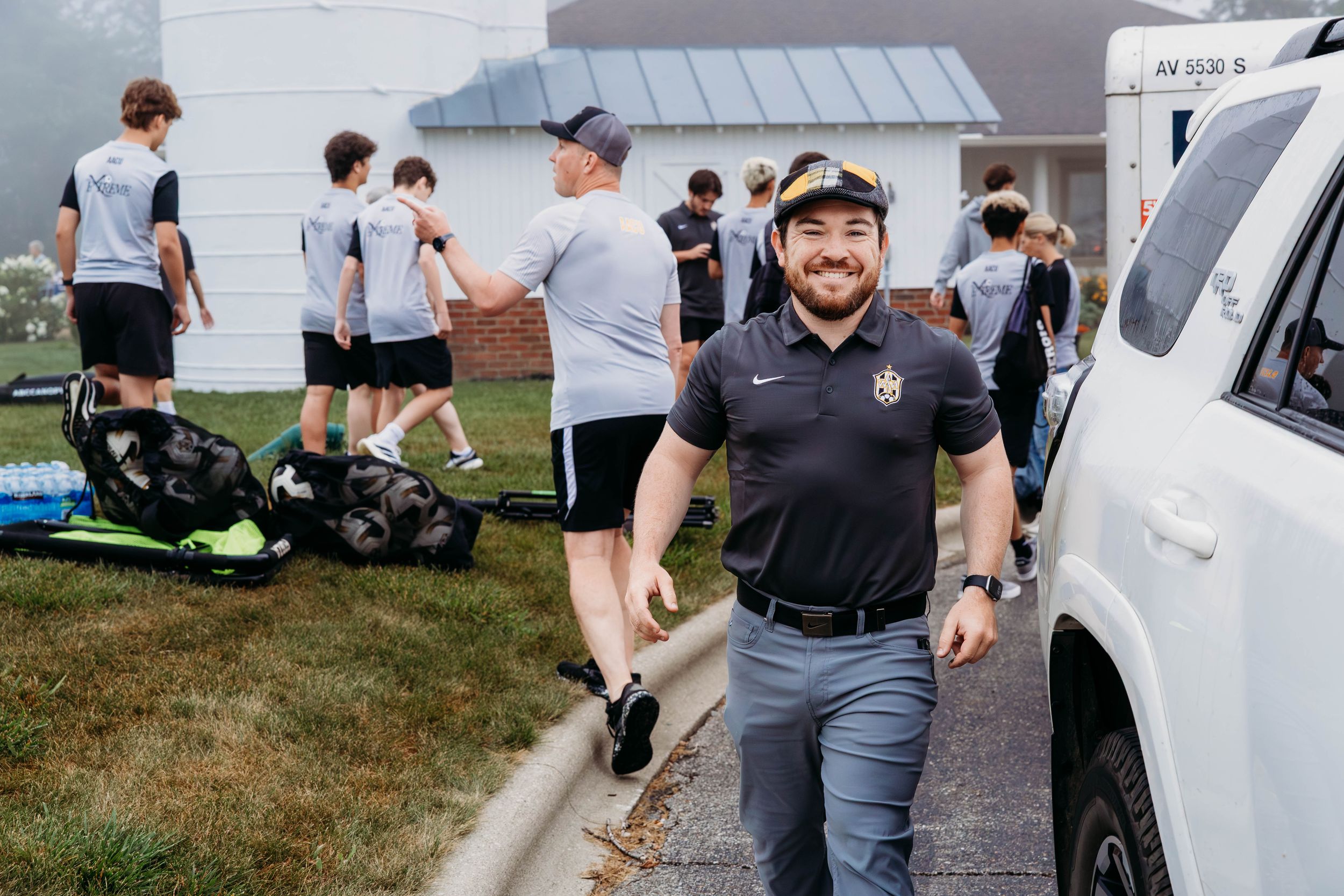 A smiling coach in a black polo walks near a white vehicle while athletes prepare equipment on a grassy field.