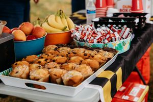 Outdoor food table with muffins, fresh peaches, bananas, and wrapped candy treats at a casual gathering event.