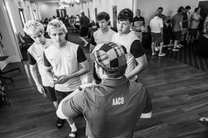 Black and white photo of youth soccer players listening to a coach in an AACU shirt during an indoor team meeting.