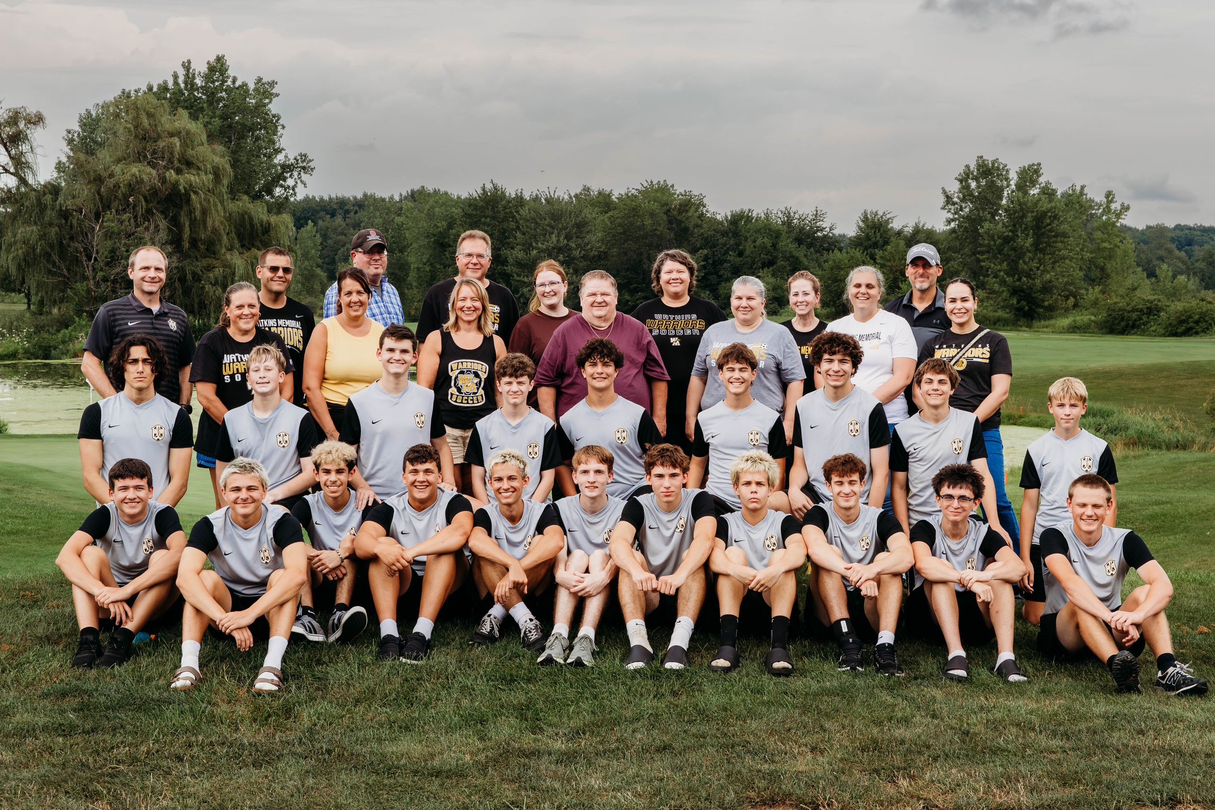Large youth sports team poses outdoors on a golf course, wearing matching black and white uniforms on a cloudy day.