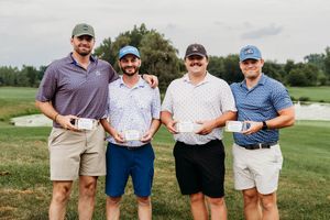 Four men holding awards pose together on a golf course after a tournament competition.