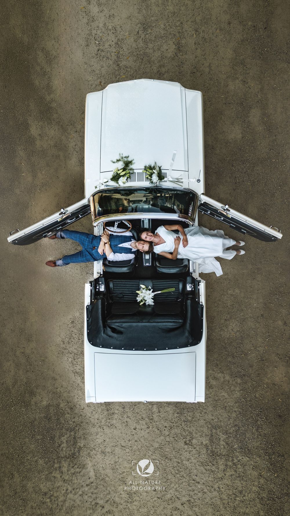 Aerial drone view of bride and groom lying in open convertible white classic wedding car, captured from above.