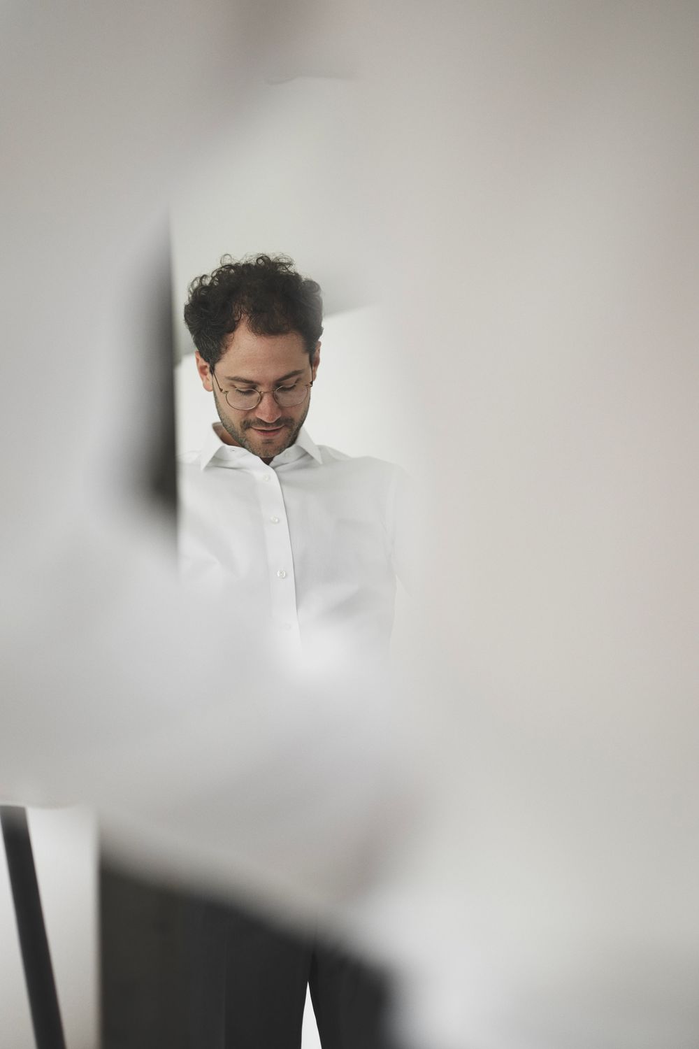 Man with curly hair and glasses wearing a white shirt, viewed through a gap in white walls.