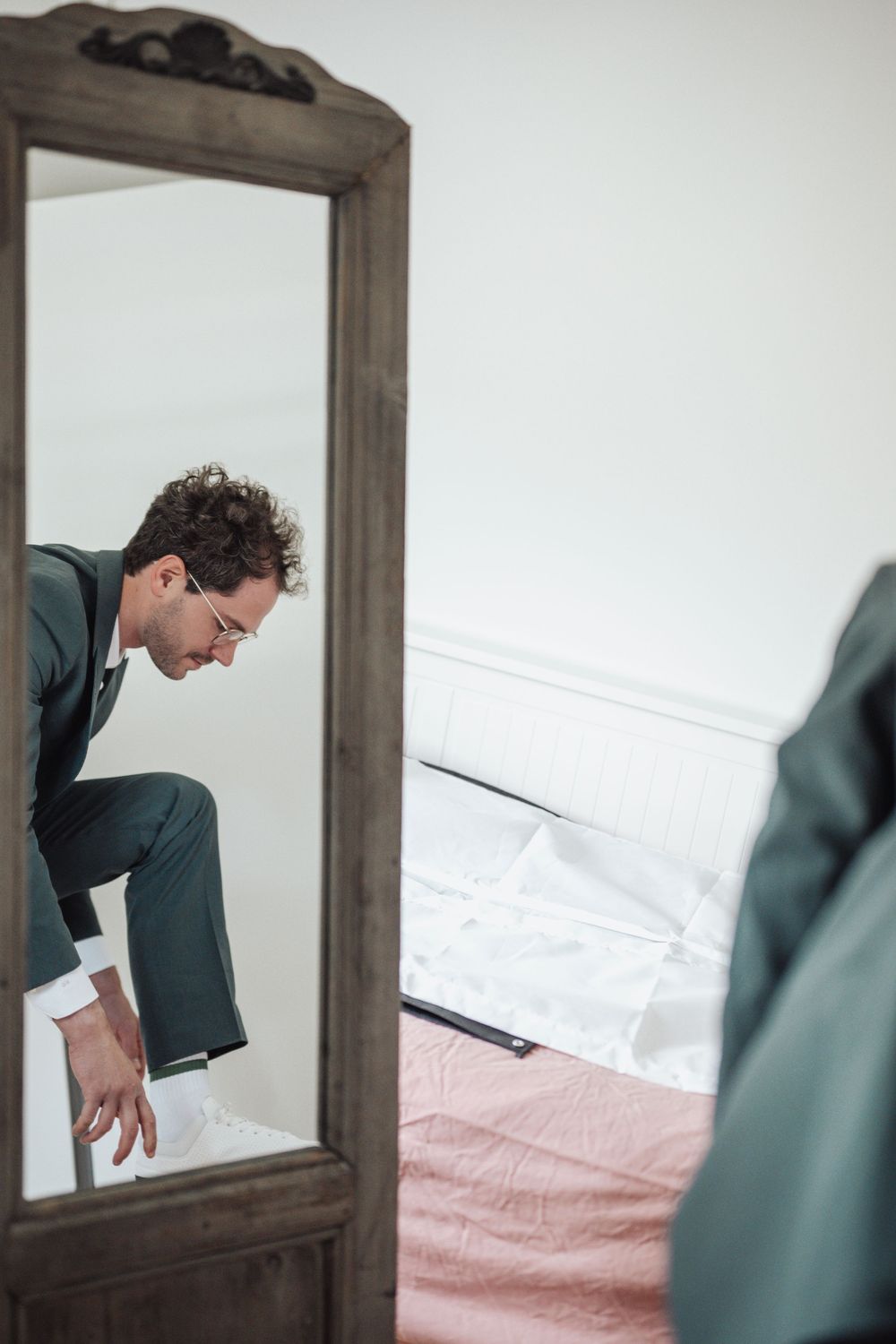 Man in green suit adjusting outfit, reflected in a wooden floor mirror beside a bed.