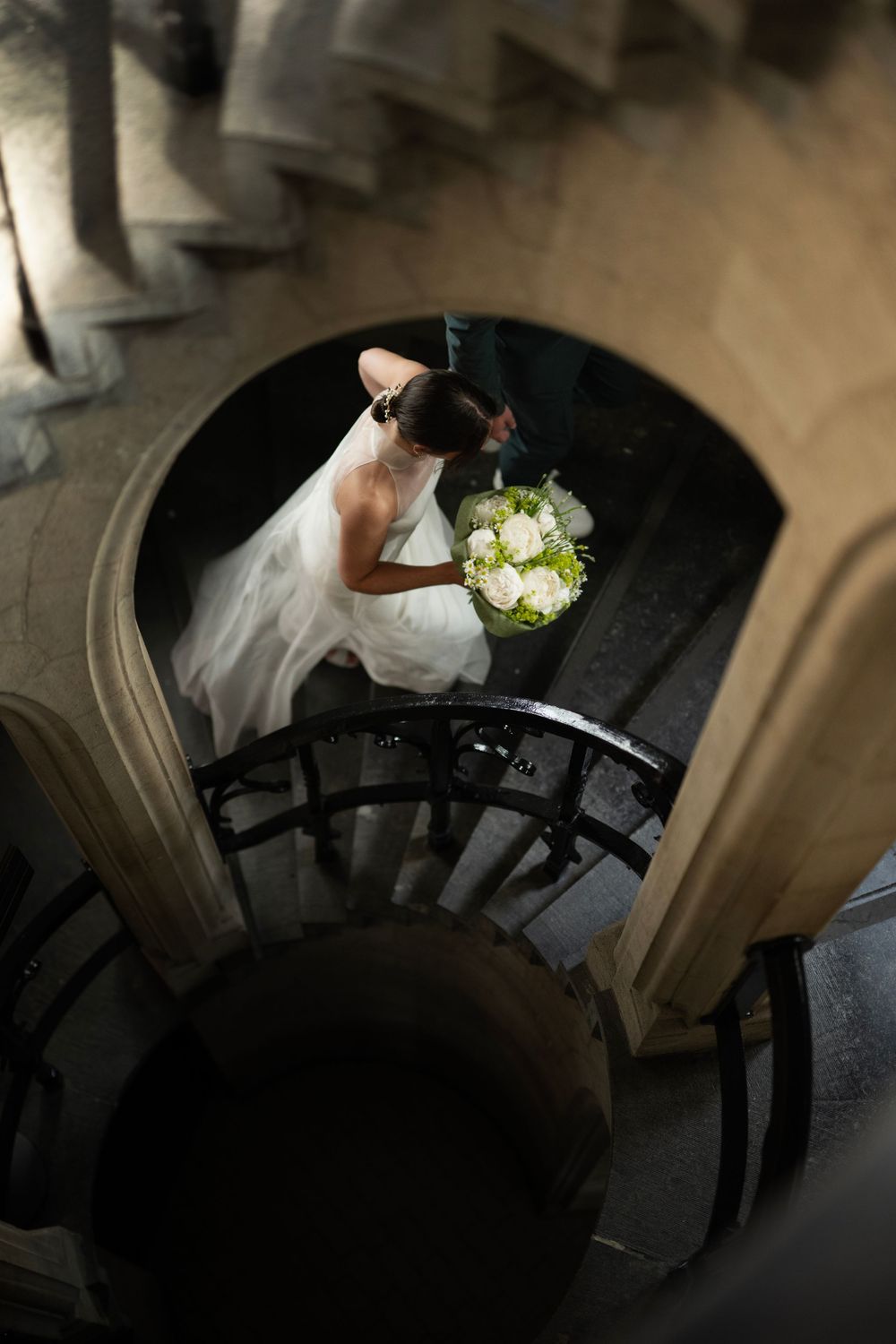 Bride in white gown holding bouquet descends elegant spiral staircase, viewed from above.