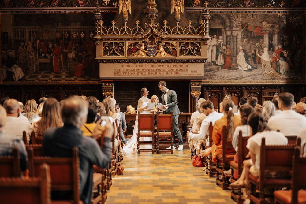 Elegant wedding ceremony in ornate historic hall with guests seated in wooden pews watching couple at altar.