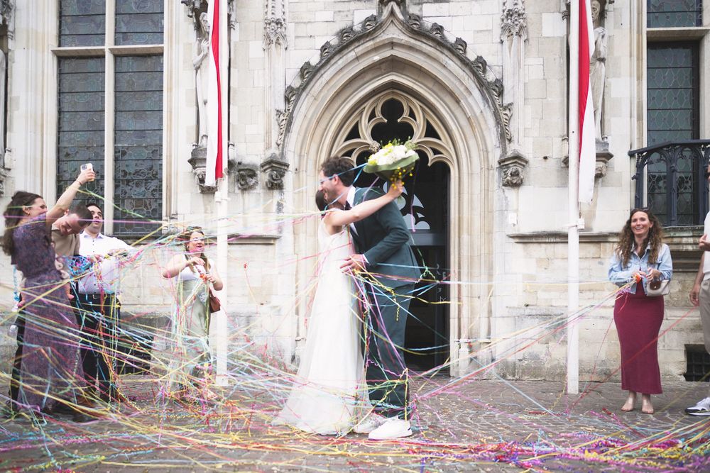 Newlyweds kiss outside gothic church entrance as guests celebrate with colorful streamers at outdoor wedding ceremony.