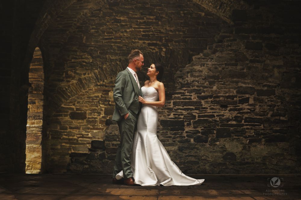 Bride and groom share a romantic moment against a rustic stone wall in a historic venue.