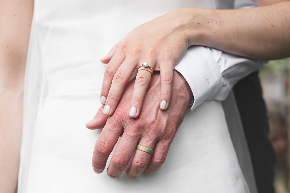 Close-up of newlywed couple hands showing wedding rings, bride in white dress embracing groom in white shirt.