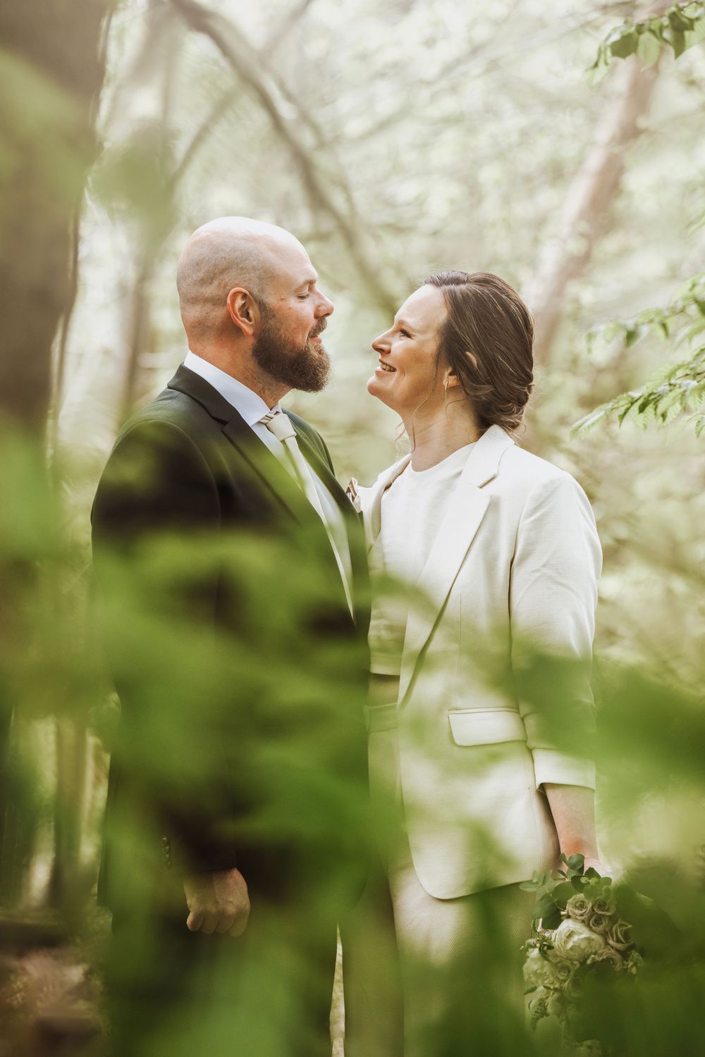 A couple in wedding attire smiles at each other surrounded by lush green foliage in a forest setting.