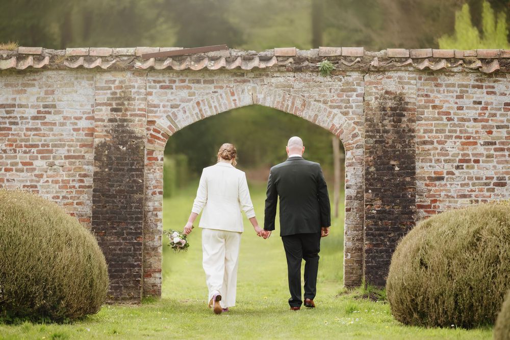 Bride and groom walking hand in hand through a rustic brick archway in a lush garden setting.