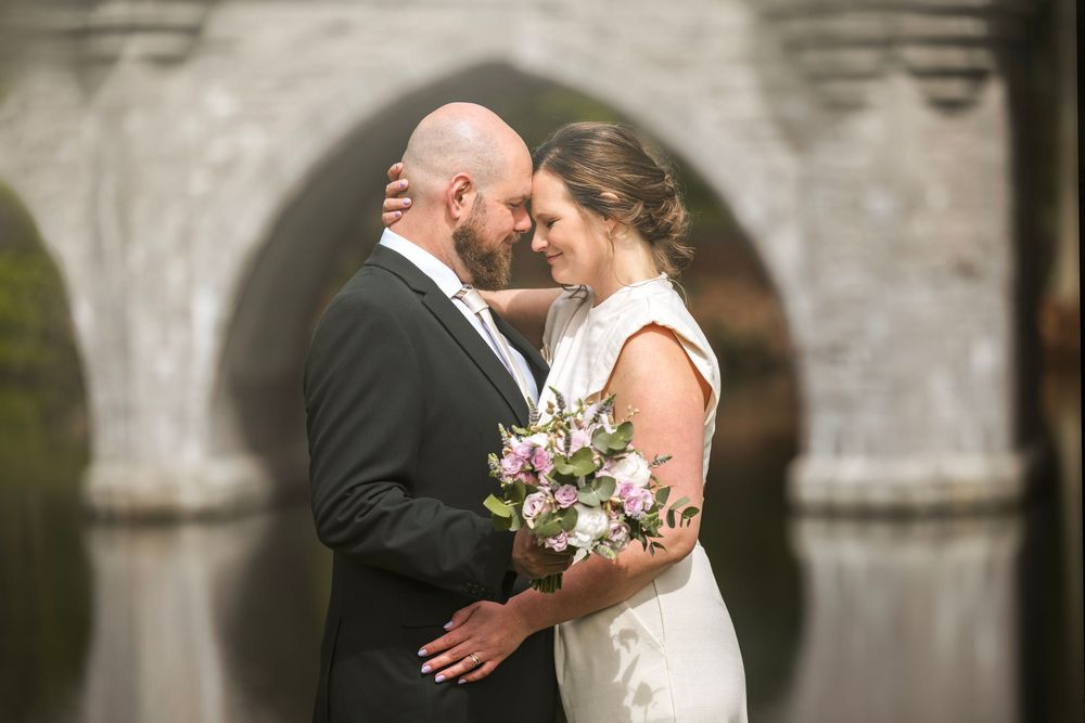 Bride and groom share a romantic moment holding a floral bouquet with an arched stone bridge reflected in water behind them.