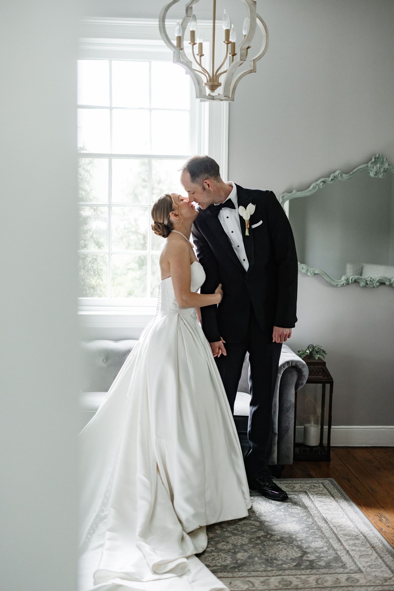 Bride and groom share a romantic kiss near a bright window inside an elegantly decorated room with chandelier.